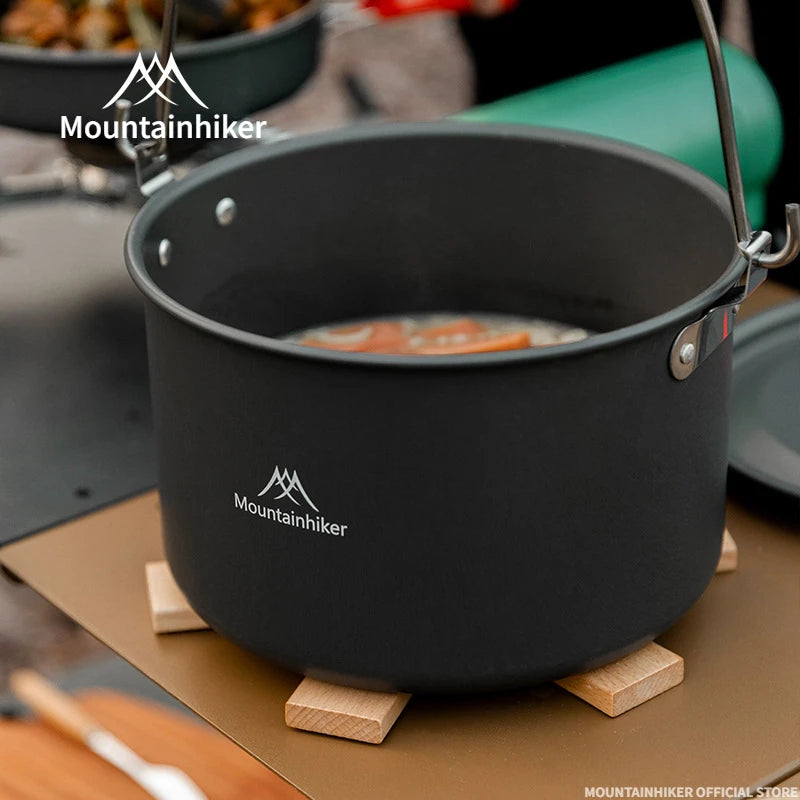 Black camping pot with Mountainhiker logo on a wooden stand, surrounded by outdoor cooking equipment.